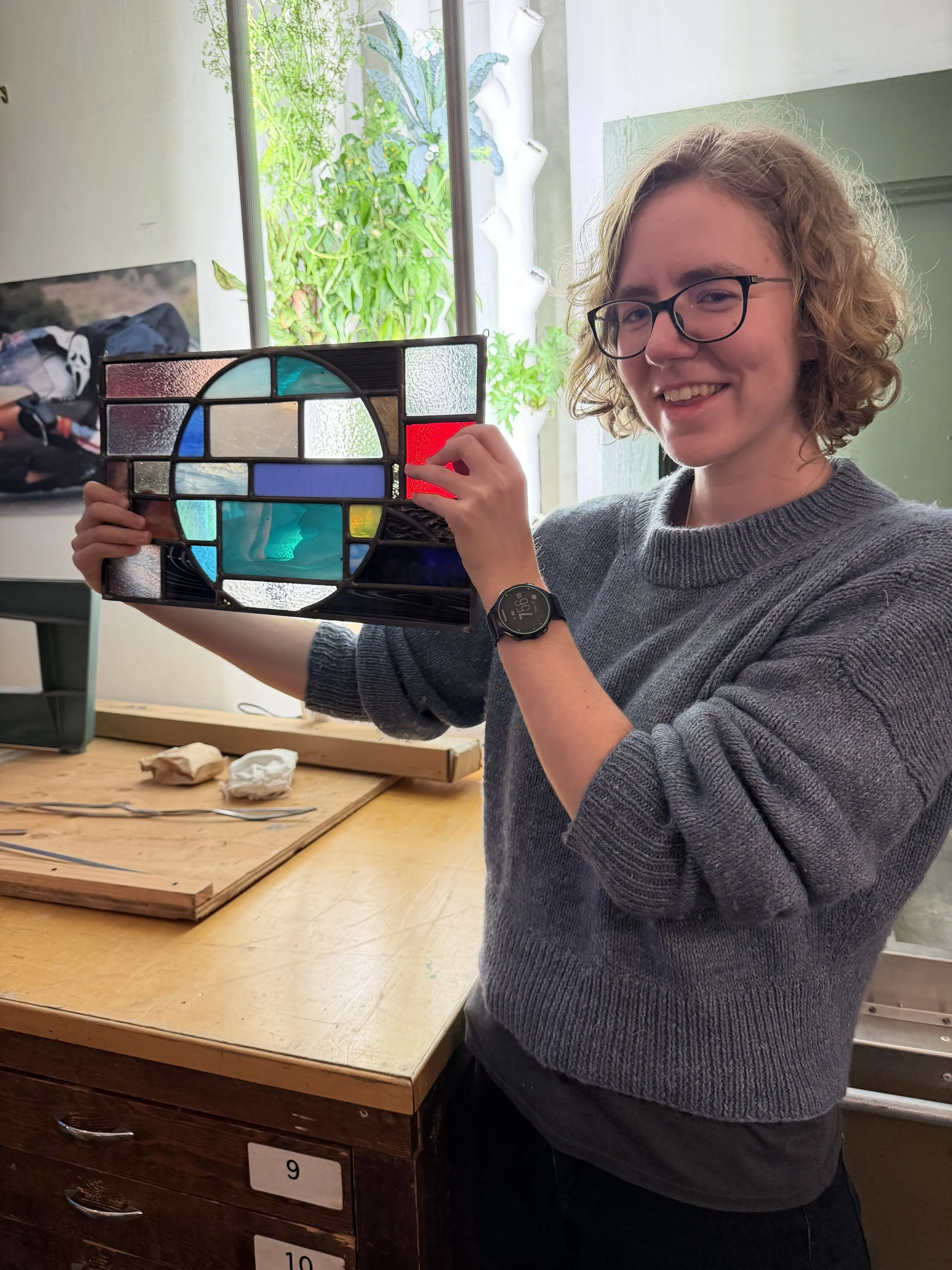 A person with curly hair and glasses smiling while holding a stained glass panel with geometric and circular designs in various colors in a room with a wooden table, plants outside a large window, and drawers numbered 9 and 10.
