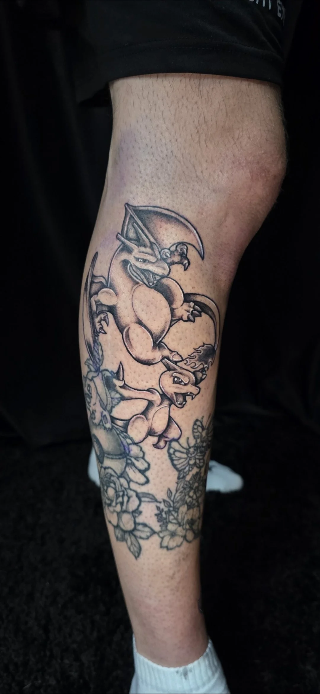Tattoo of two Pokémon characters, Charizard and Blaziken, on person's leg with floral design below.