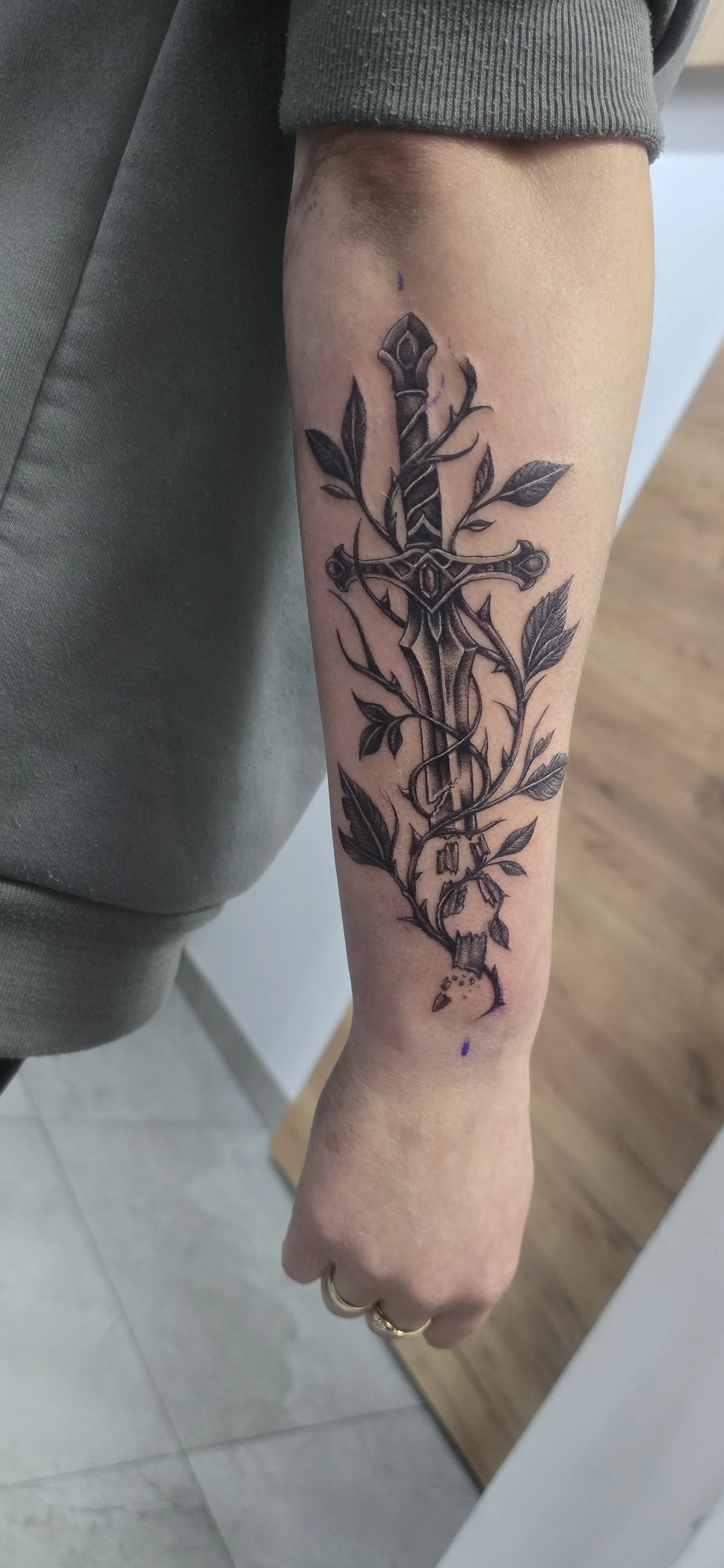 Black and gray tattoo of a sword entwined with vines and leaves on a person's forearm.