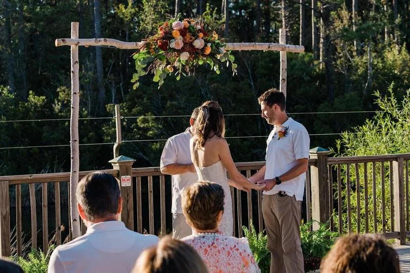 A couple exchanging vows at an outdoor wedding ceremony on a wooden deck with a floral arch, surrounded by trees and sunlight.