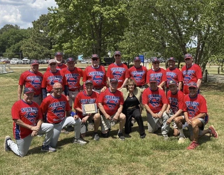 A group of middle-aged and older adults posing for a photo on a baseball field, wearing green jerseys and caps with yellow lettering that read 'Charlottesville Retreads.' They are standing and kneeling on the dirt infield, surrounded by a chain-link fence and trees in the background.