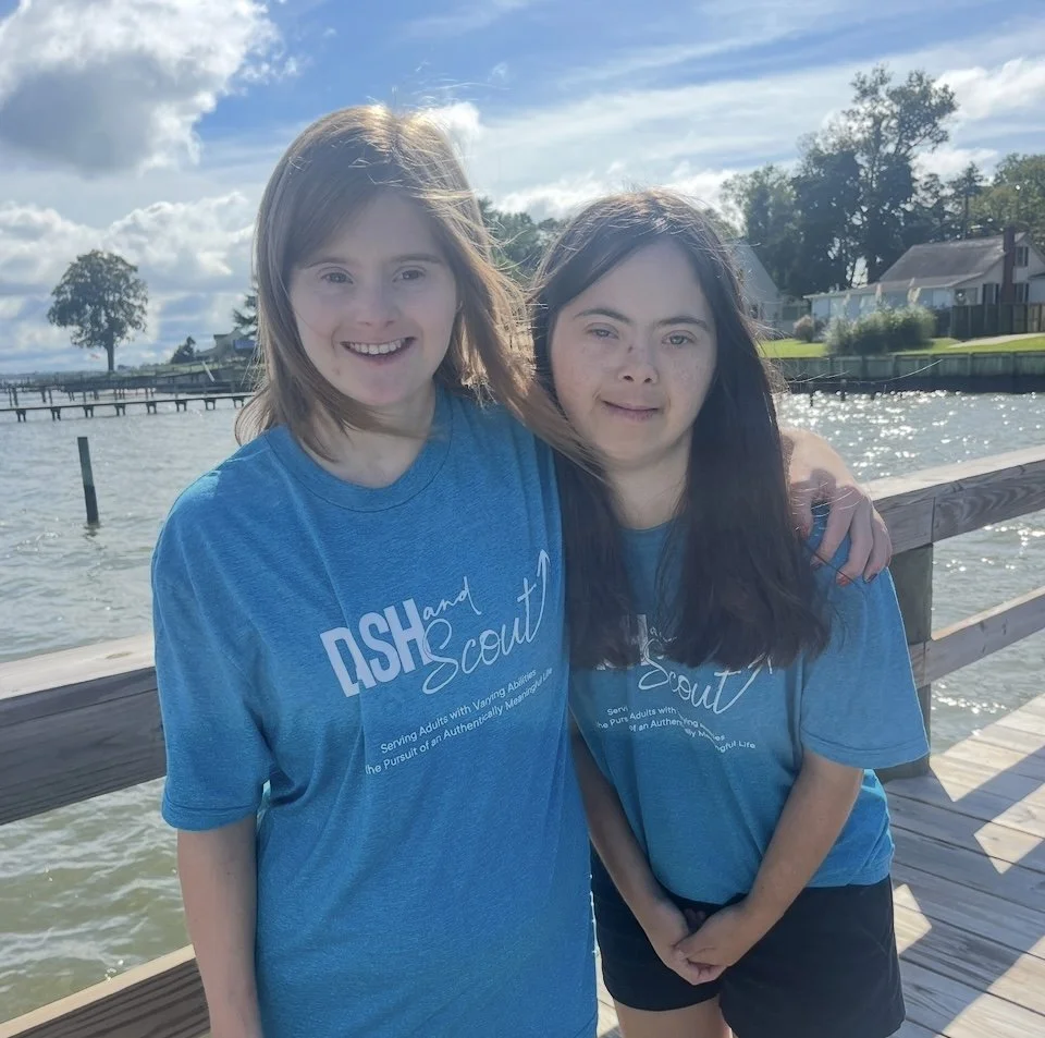 Two young women standing on a wooden pier near a body of water, smiling, with trees, houses, and a partly cloudy sky in the background, wearing matching blue t-shirts.