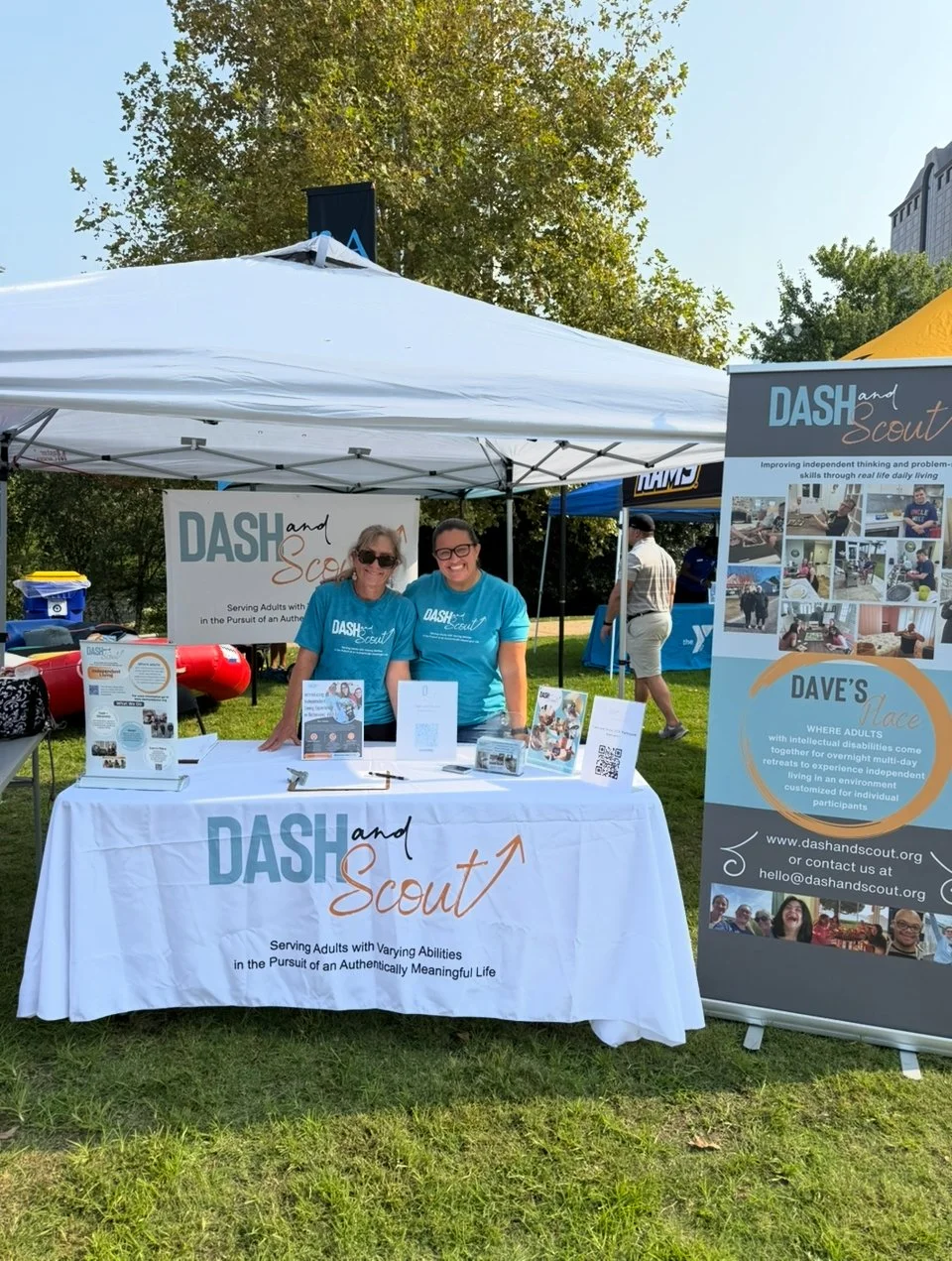 Two women smiling at a booth under a white canopy tent, promoting DASH and Scout organization at an outdoor event. The booth has a table with informational materials and a large banner on a stand beside it.