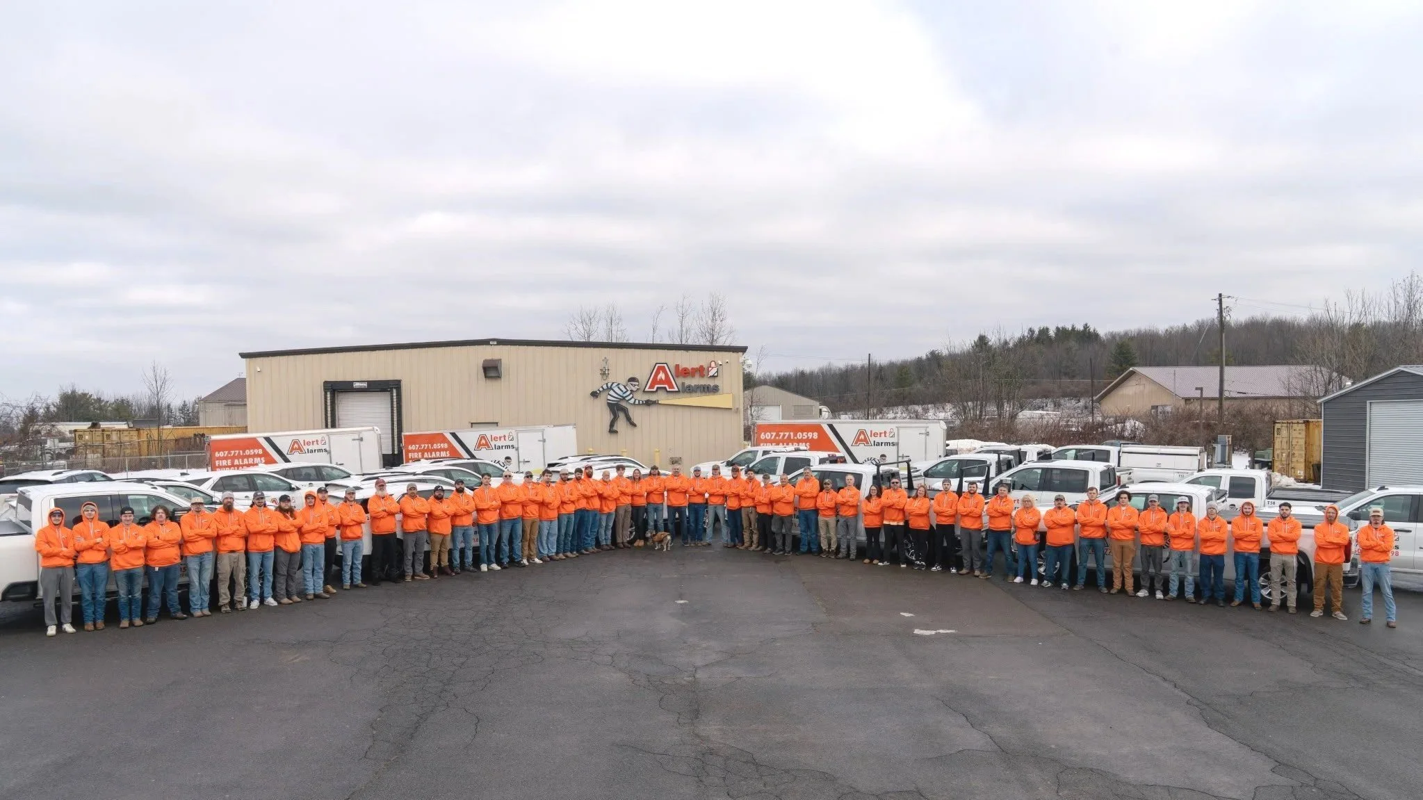 Group of workers in orange jackets standing in a line outside a building with alarm company signage and vehicles around.