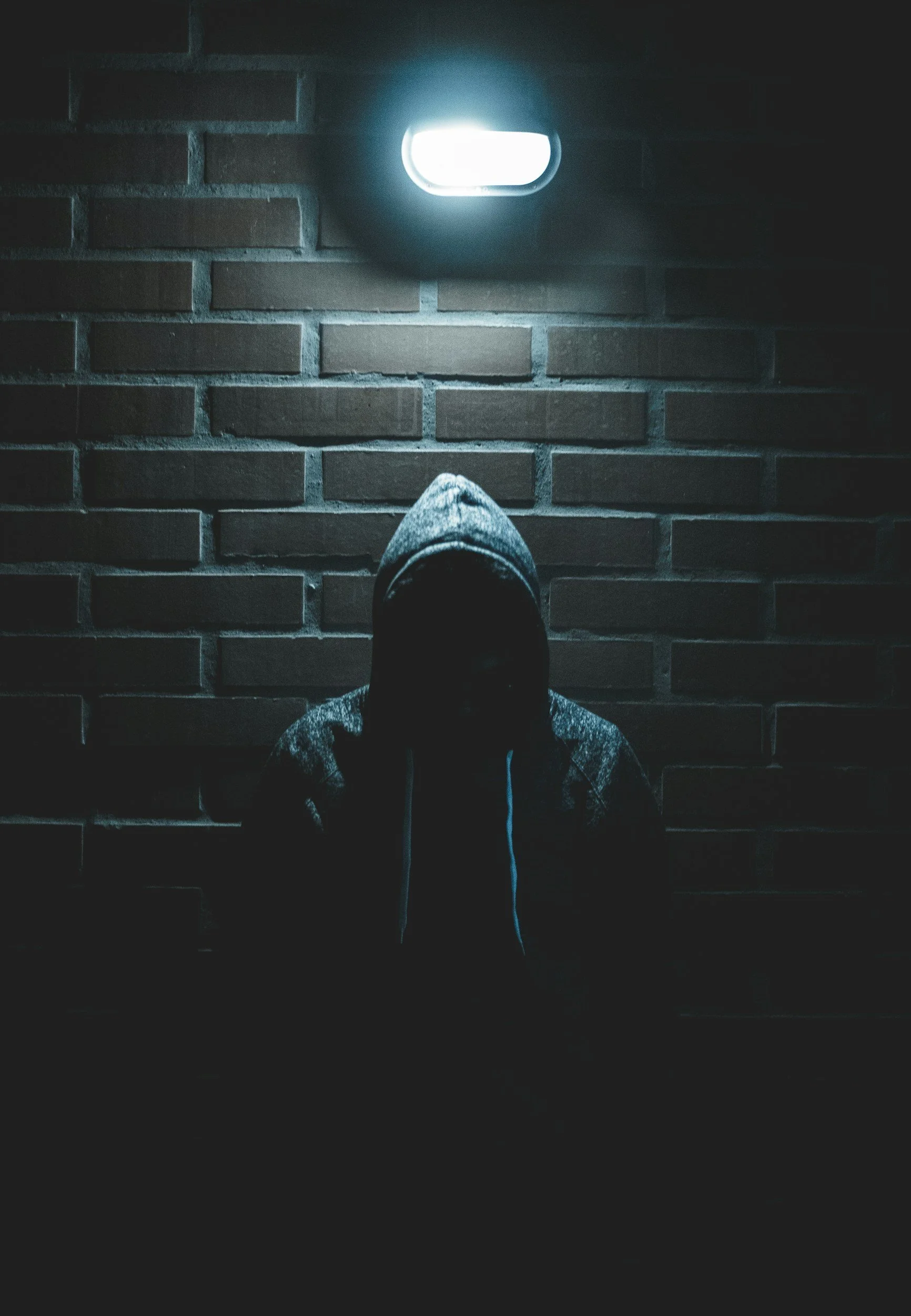 A person wearing a hoodie with the hood up, sitting against a brick wall, partially in darkness with a bright light above.