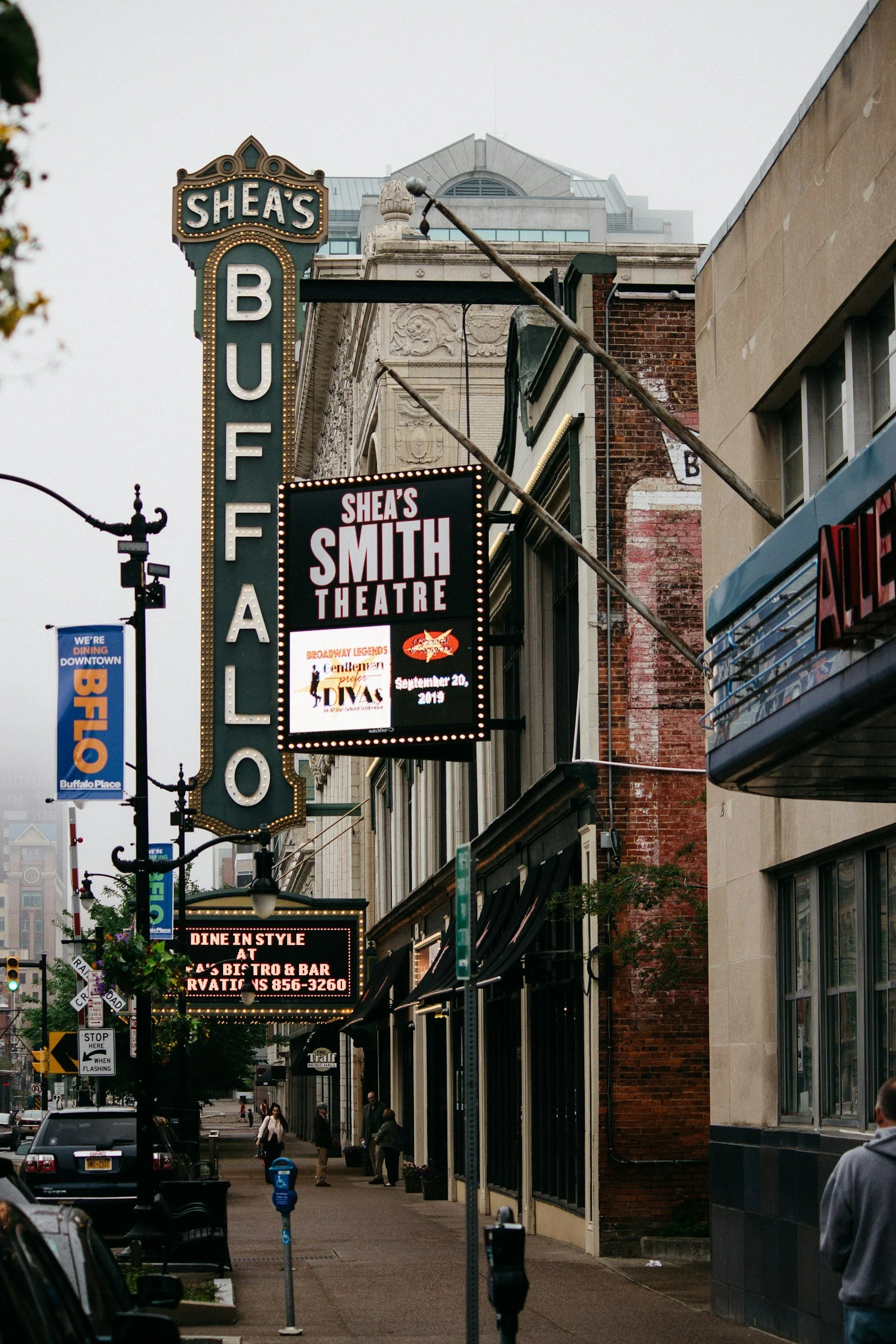 Street view of Shea's Buffalo Theatre with illuminated marquee and signage, showing upcoming show dates and details, surrounded by other downtown buildings and pedestrians.