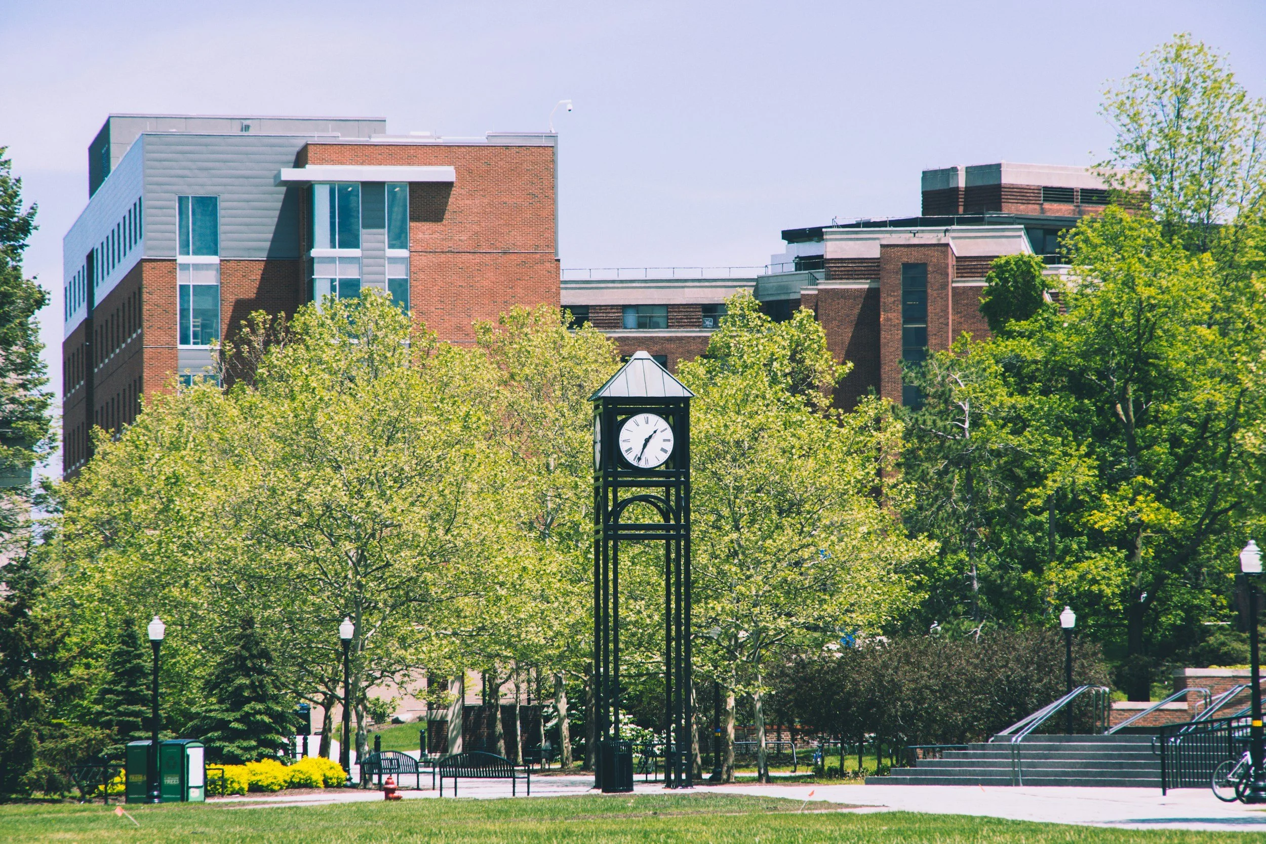 A park with a clock tower, green trees, benches, street lamps, and a backdrop of modern brick and glass buildings.