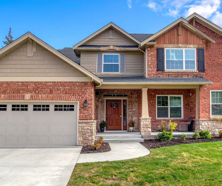 Front view of a two-story house with a brick facade, gray siding, and a peaked roof. It has a closed garage door, a front door with a small window, and a porch with potted plants. The lawn is green with some freshly planted bushes. The sky is blue with some clouds.