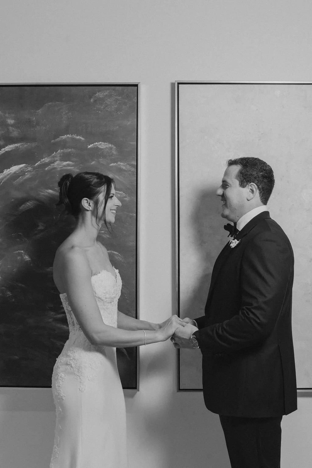 A black-and-white photo of a bride and groom holding hands and smiling at each other during their wedding, standing in front of framed artwork on a wall.