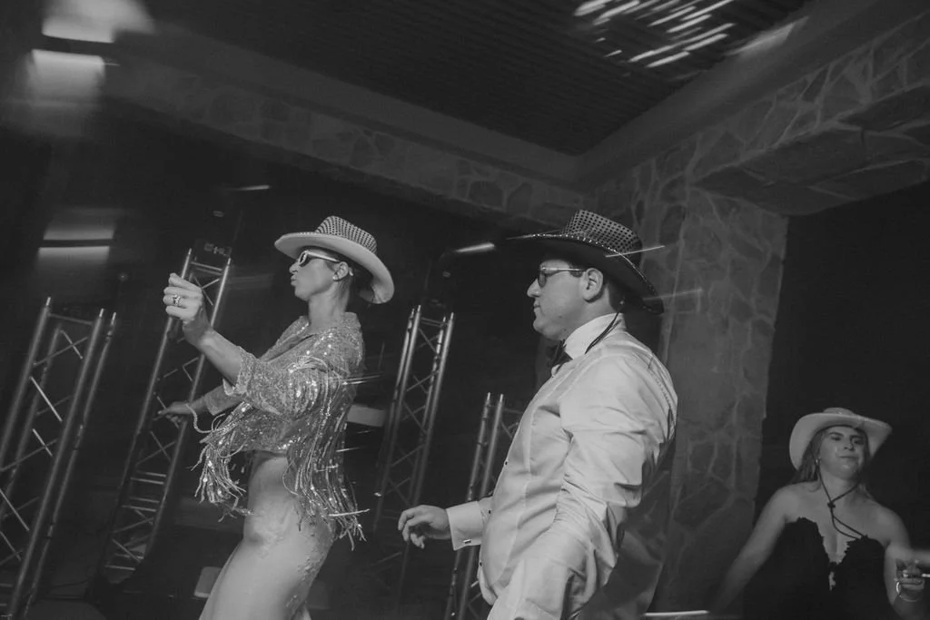 Black and white photo of three people wearing cowboy hats, dressed in glamorous outfits, at a party or performance, with metal truss structures in the background.