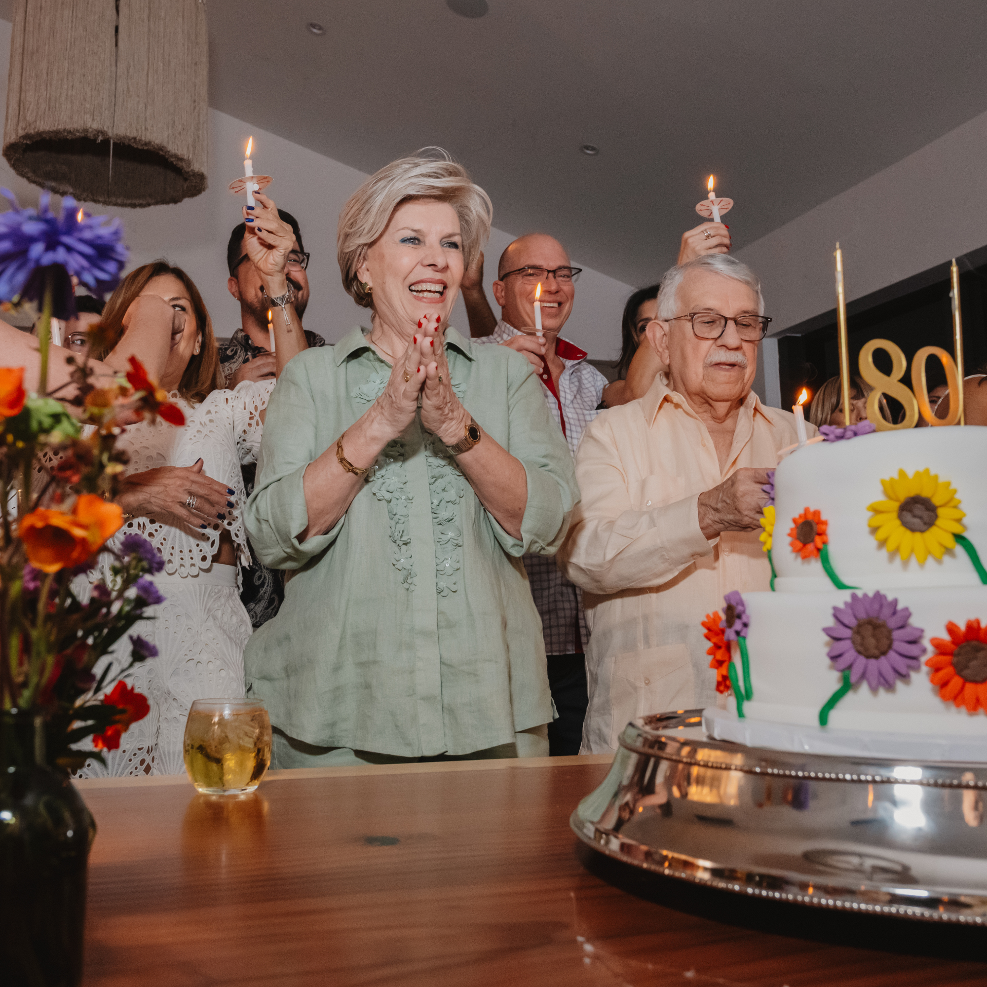 A group of people celebrating an 80th birthday with a decorated cake featuring colorful flowers and a number 80 topper. The woman at the center is clapping and smiling, and others are holding lit candles.