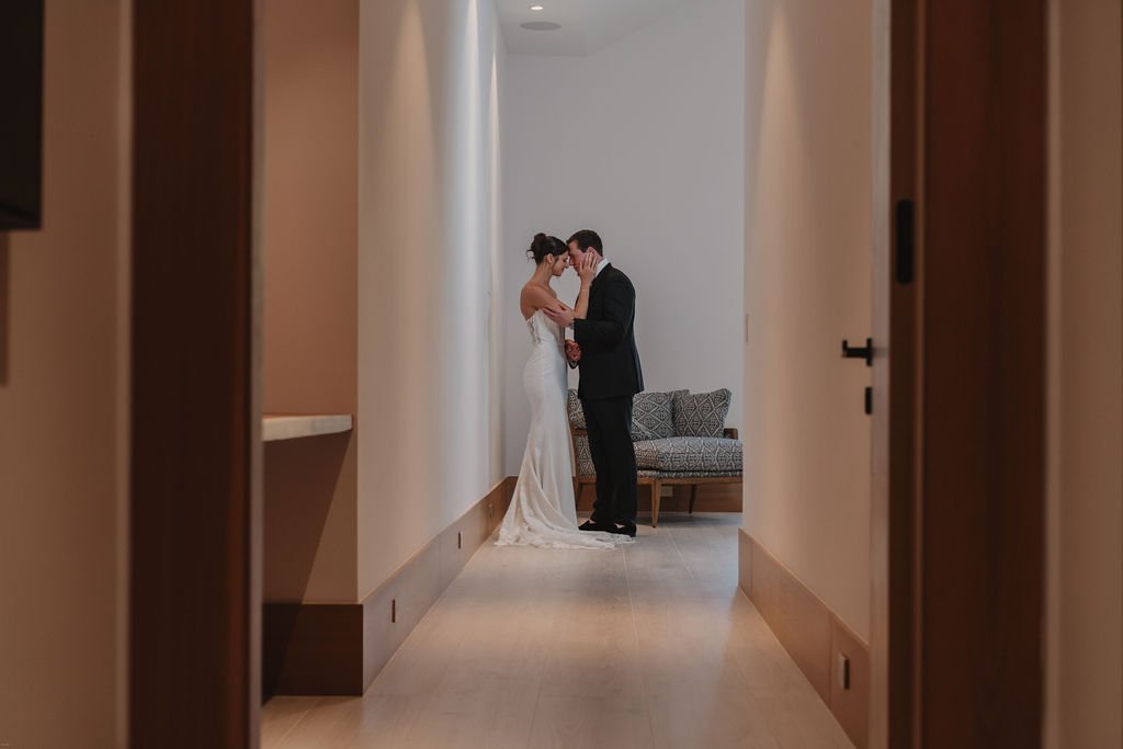 A bride and groom standing close together in a hallway, sharing an intimate moment on their wedding day.