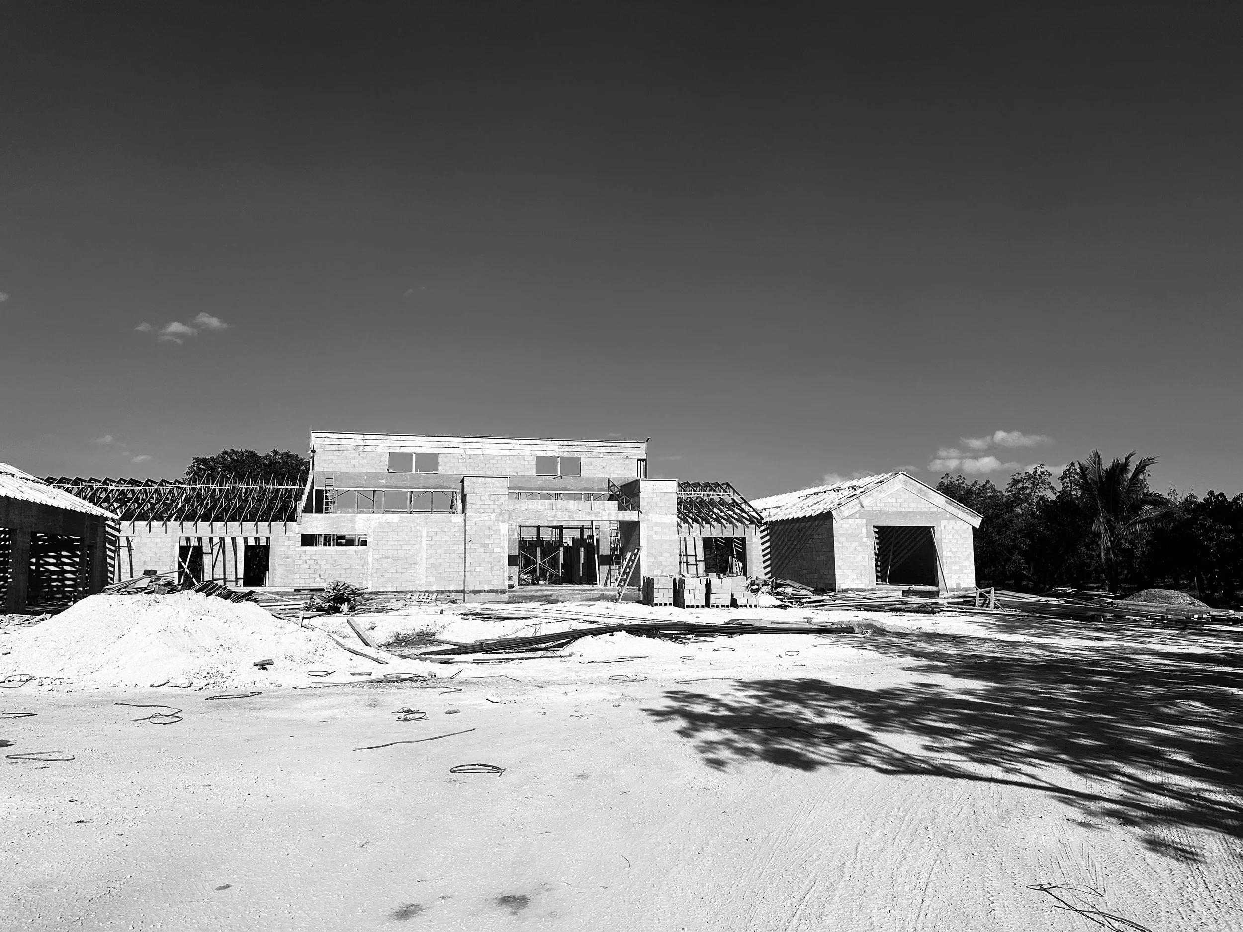 Under construction building with two visible structures and construction materials on a dirt site, surrounded by trees, in black and white.