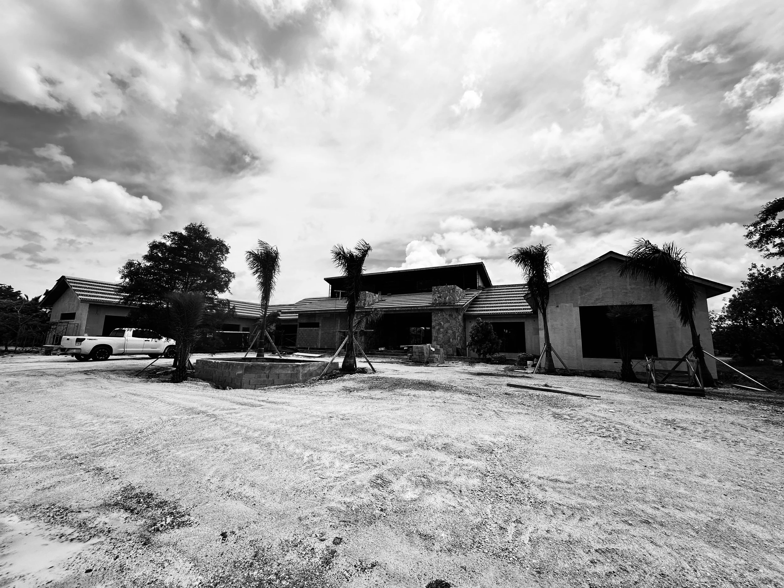 Black and white photo of a house under construction with palm trees in the front yard and a pickup truck parked to the left outside. The sky is filled with clouds.