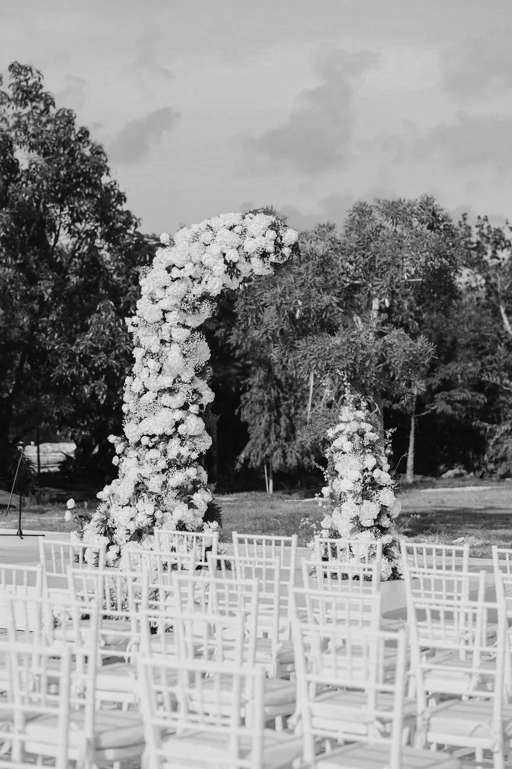 Outdoor wedding setup with a floral arch and white chairs in a natural setting.