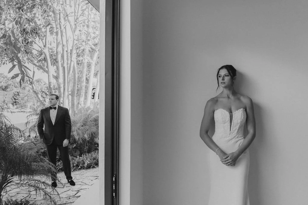 A black and white photo of a bride in a strapless wedding gown standing against a plain wall, looking to the right, with her hands clasped in front. Outside the window, a groom in a tuxedo stands outdoors among trees and plants, looking straight ahea