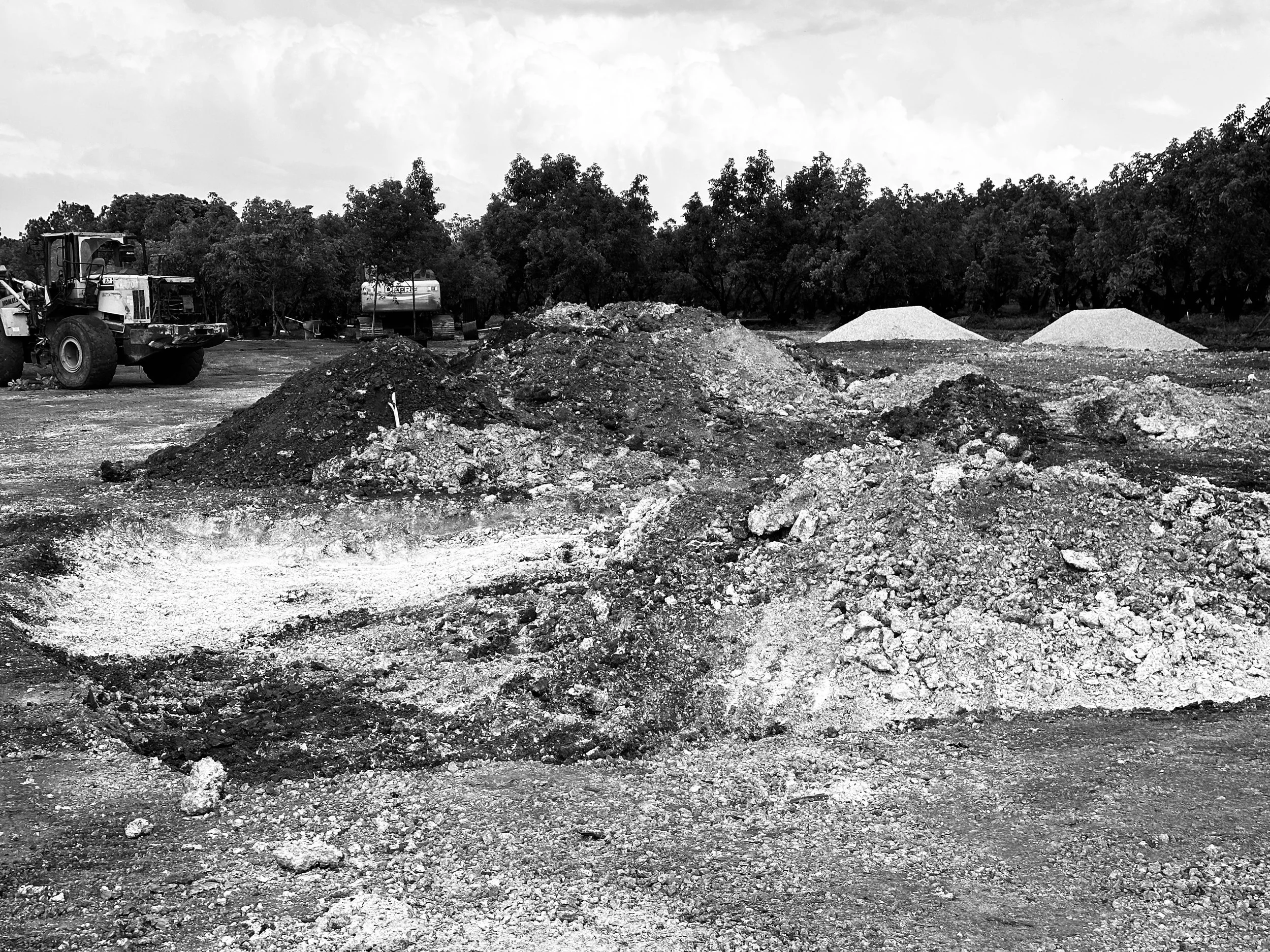 Construction site with piles of dirt, machinery, and gravel mounds with trees in the background.