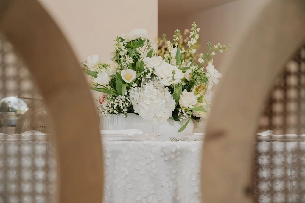 Elegant table setting with a large white floral arrangement, including white roses, hydrangeas, and greenery, partially viewed through a wooden chair back.