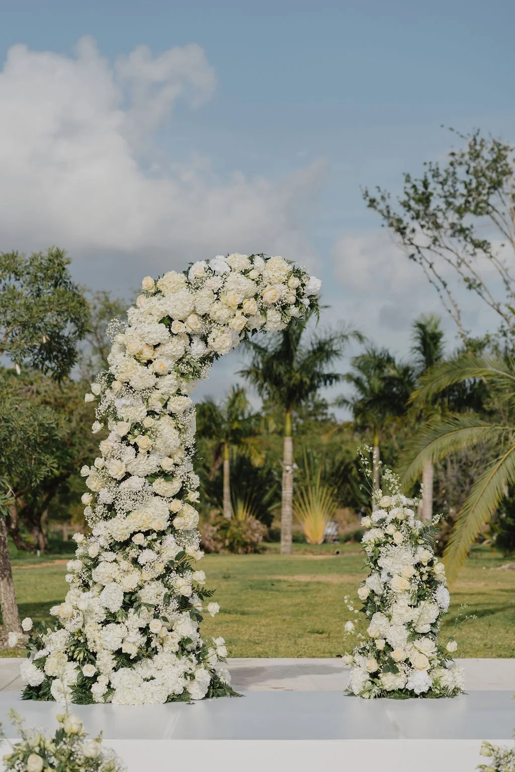Wedding arch decorated with white flowers set outdoors with trees and blue sky in the background.