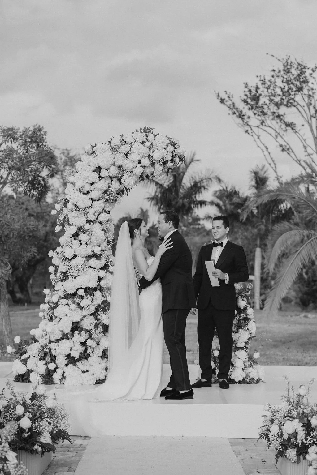 A black and white photo of a wedding ceremony outdoors with a bride and groom exchanging vows under a floral arch, with an officiant standing nearby.