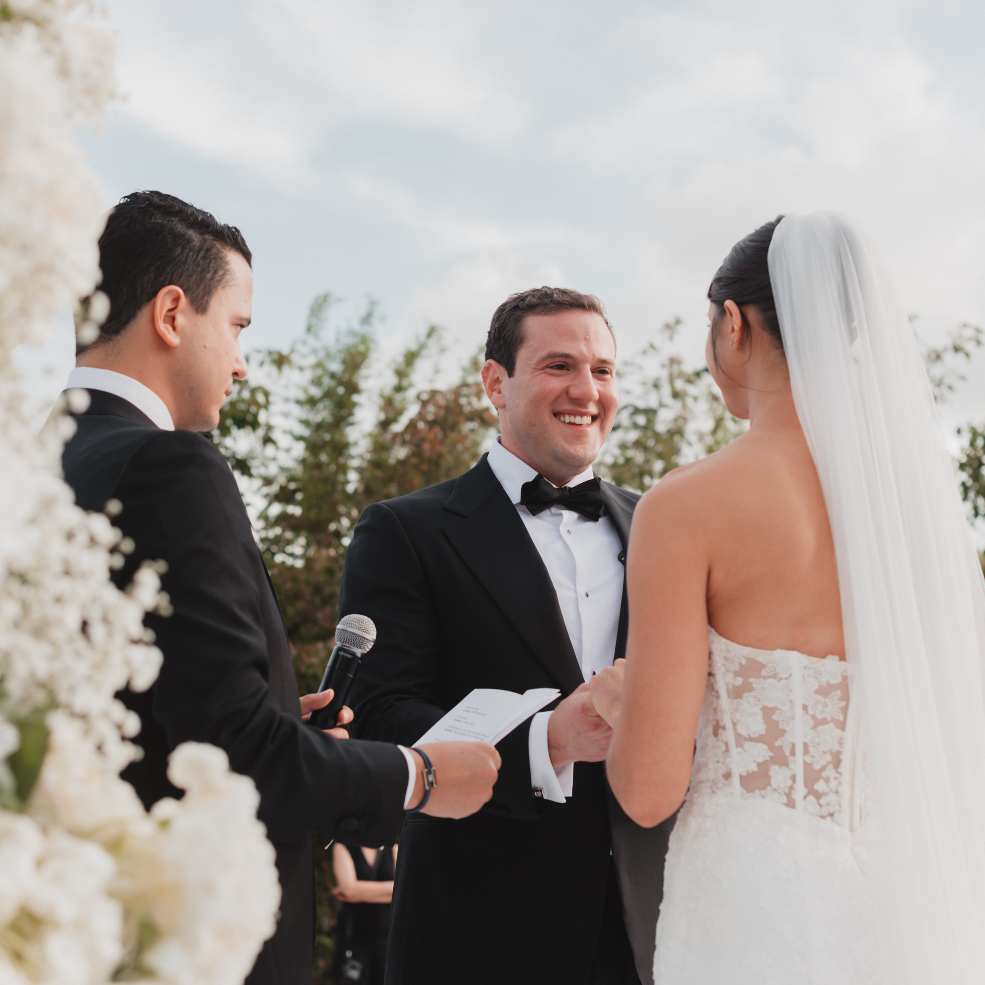 A wedding ceremony outdoors with a groom and bride exchanging vows, officiant reading from a book, and groom smiling at bride, surrounded by flowers and trees.