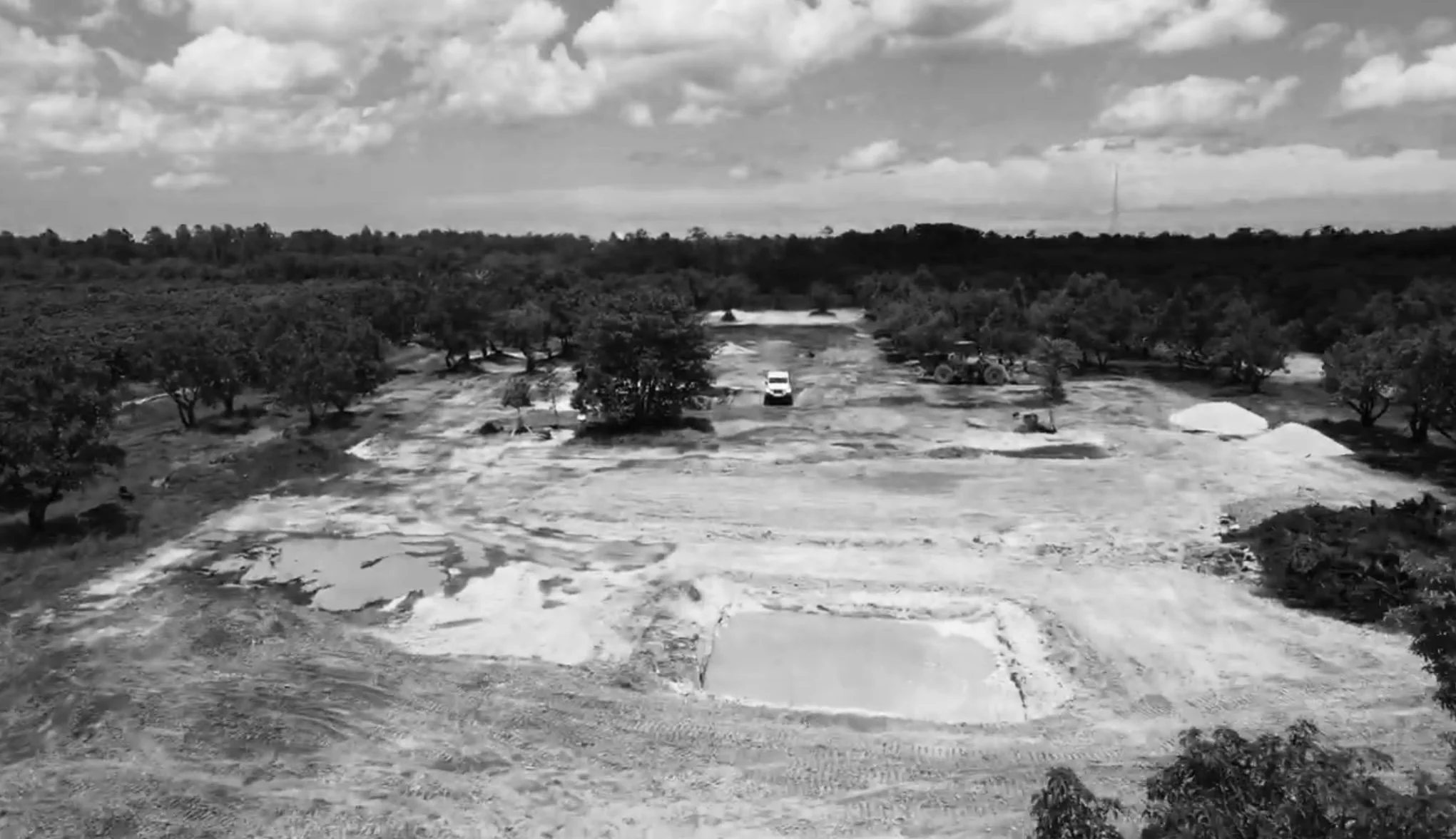 A black and white aerial view of a construction site in a rural area, with trees surrounding the site and a single vehicle present.