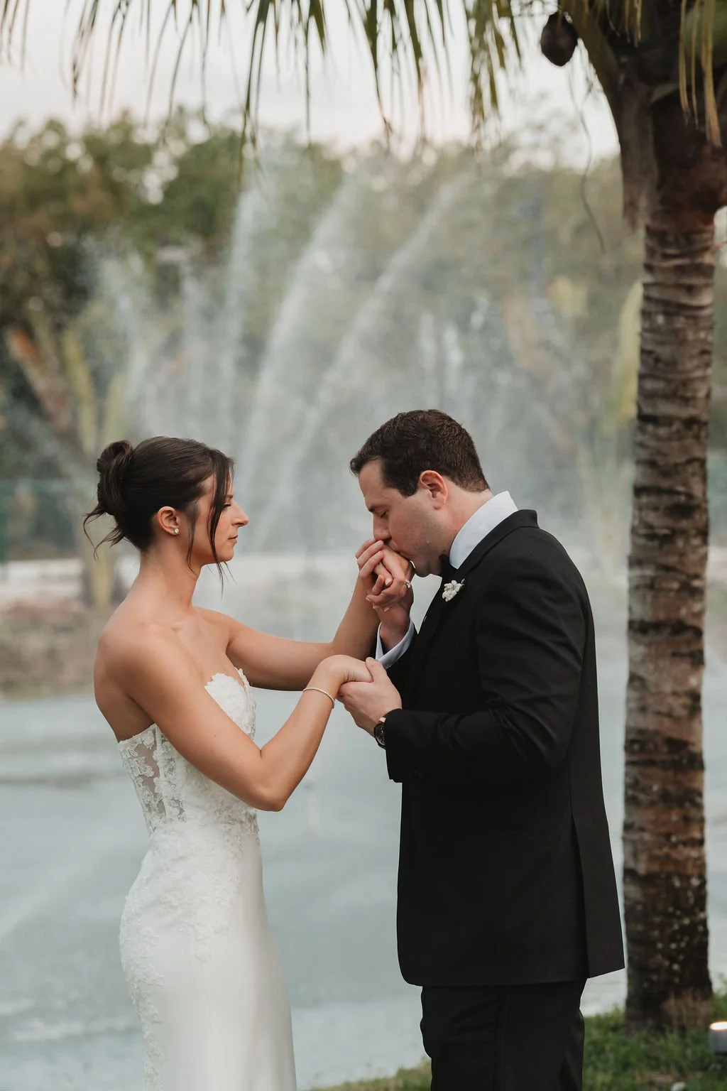 A bride and groom sharing a tender moment outdoors near water, with the groom kissing the bride's hand, surrounded by trees and palm leaves.