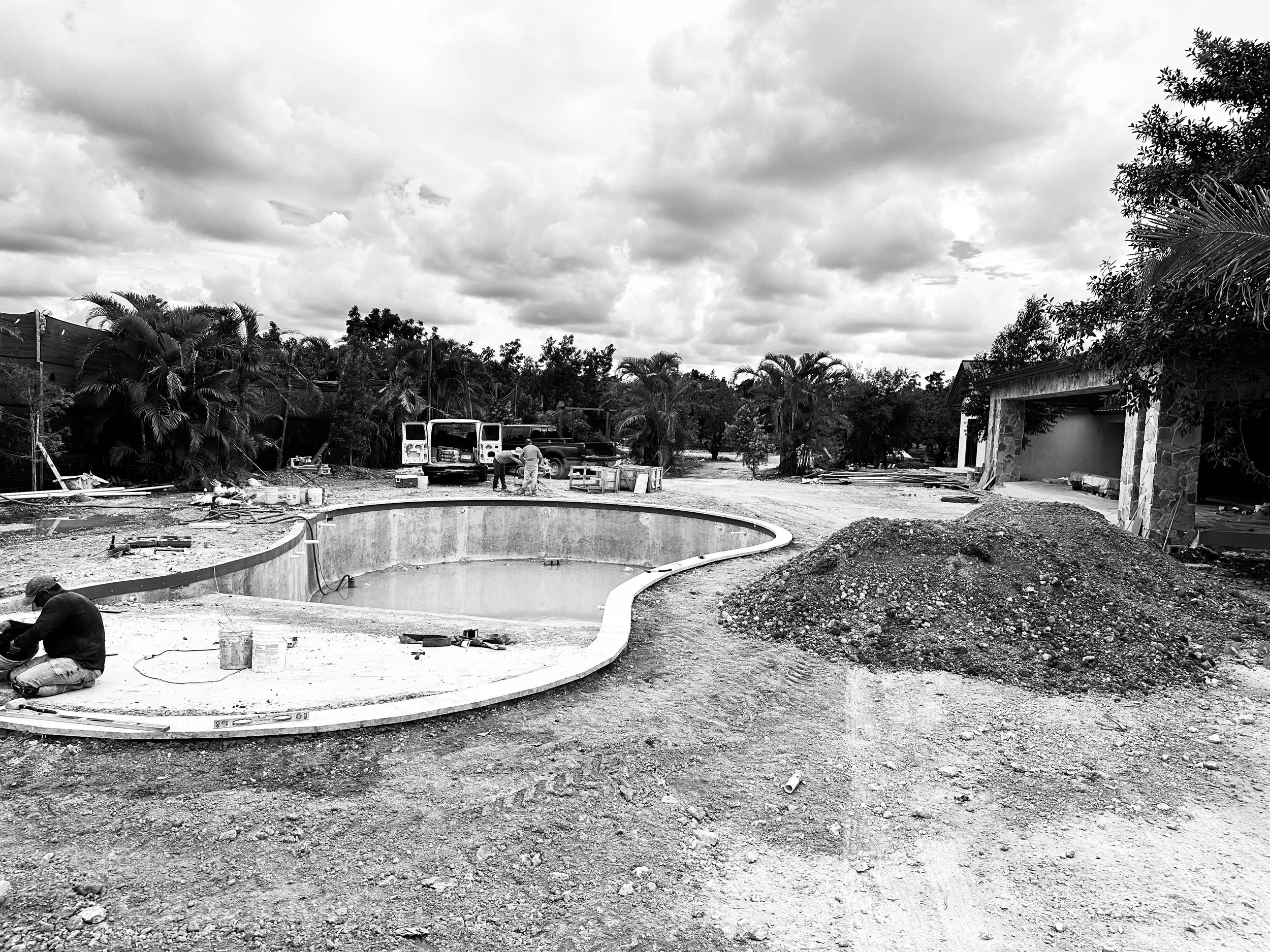 Construction site with a partially built round swimming pool, construction workers, piles of dirt, construction vehicles, and surrounding trees.