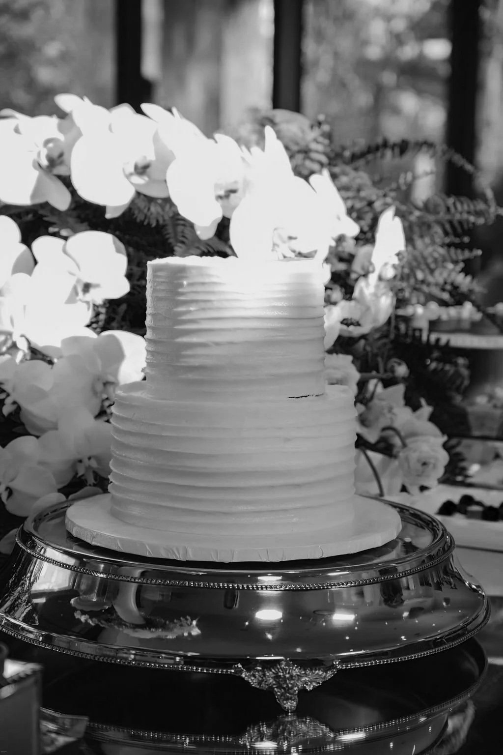 A tall, layered white cake with horizontal ridges on a cake stand, surrounded by white flowers, in black and white photo.