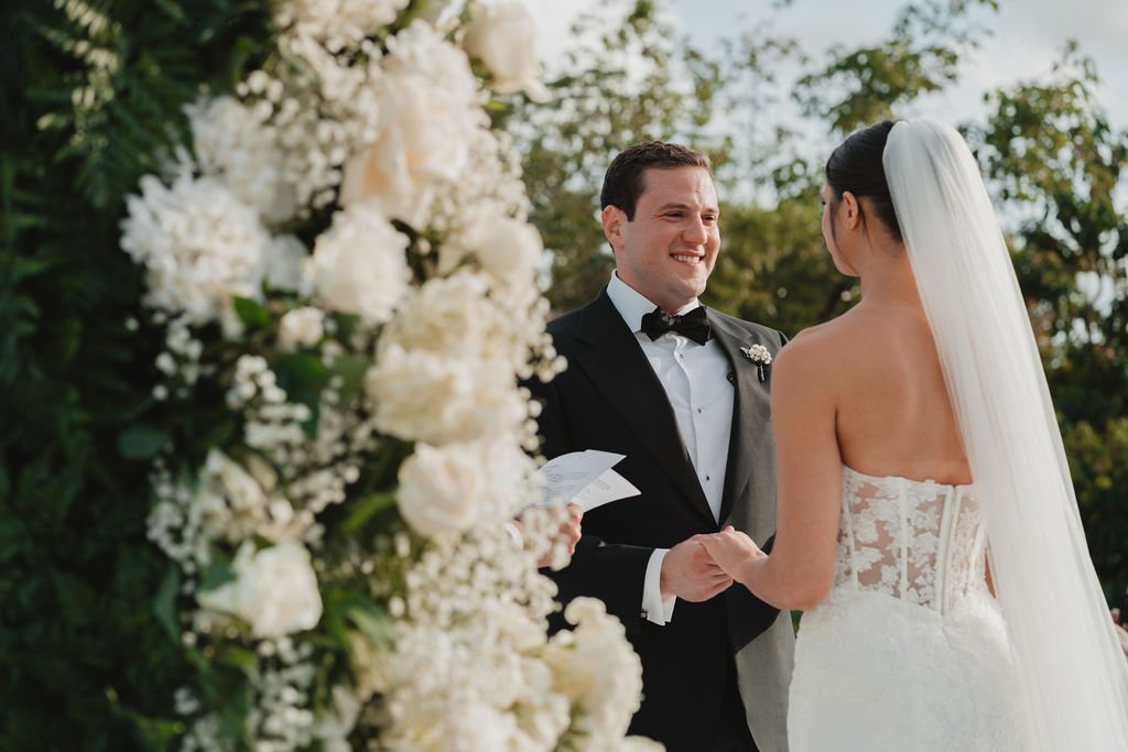 A bride and groom holding hands during their wedding ceremony outdoors, with a floral arch and greenery in the background.
