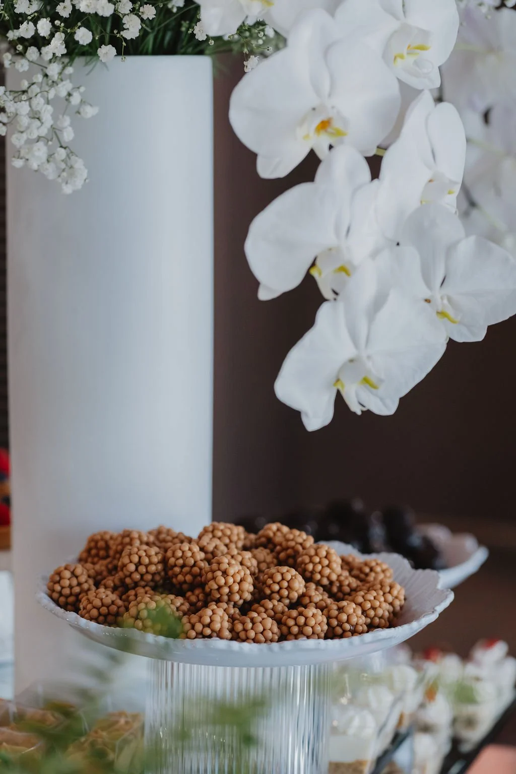 White orchids in a tall white vase with a bowl of small round brown snacks or candies in the foreground.