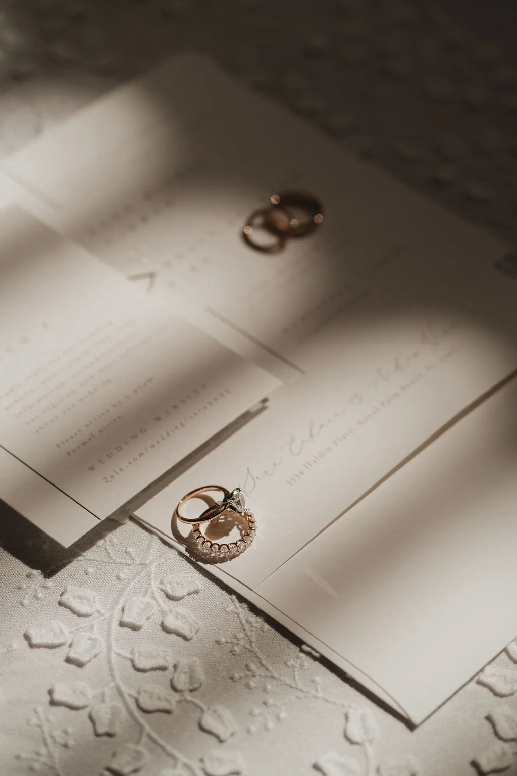 A close-up of a wedding ring with a diamond, placed on a white embroidered cloth and an invitation card, with soft lighting creating shadows and reflections.