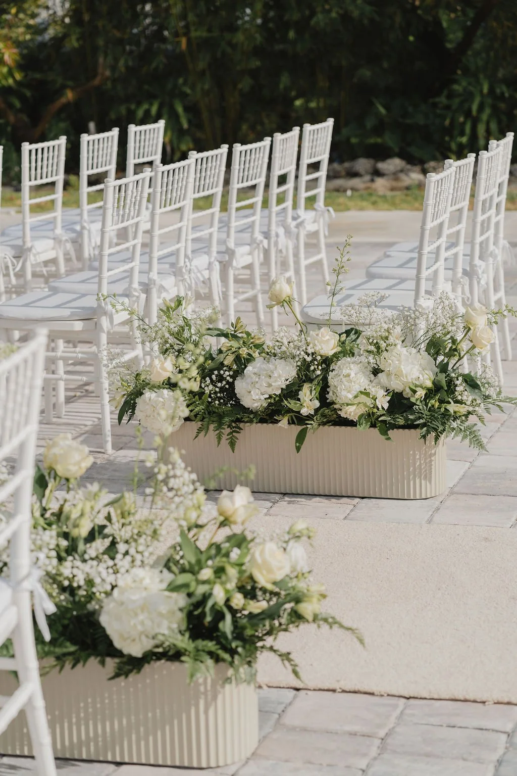White chairs arranged outdoors with large white floral arrangements in rectangular planters, set up for a wedding ceremony.