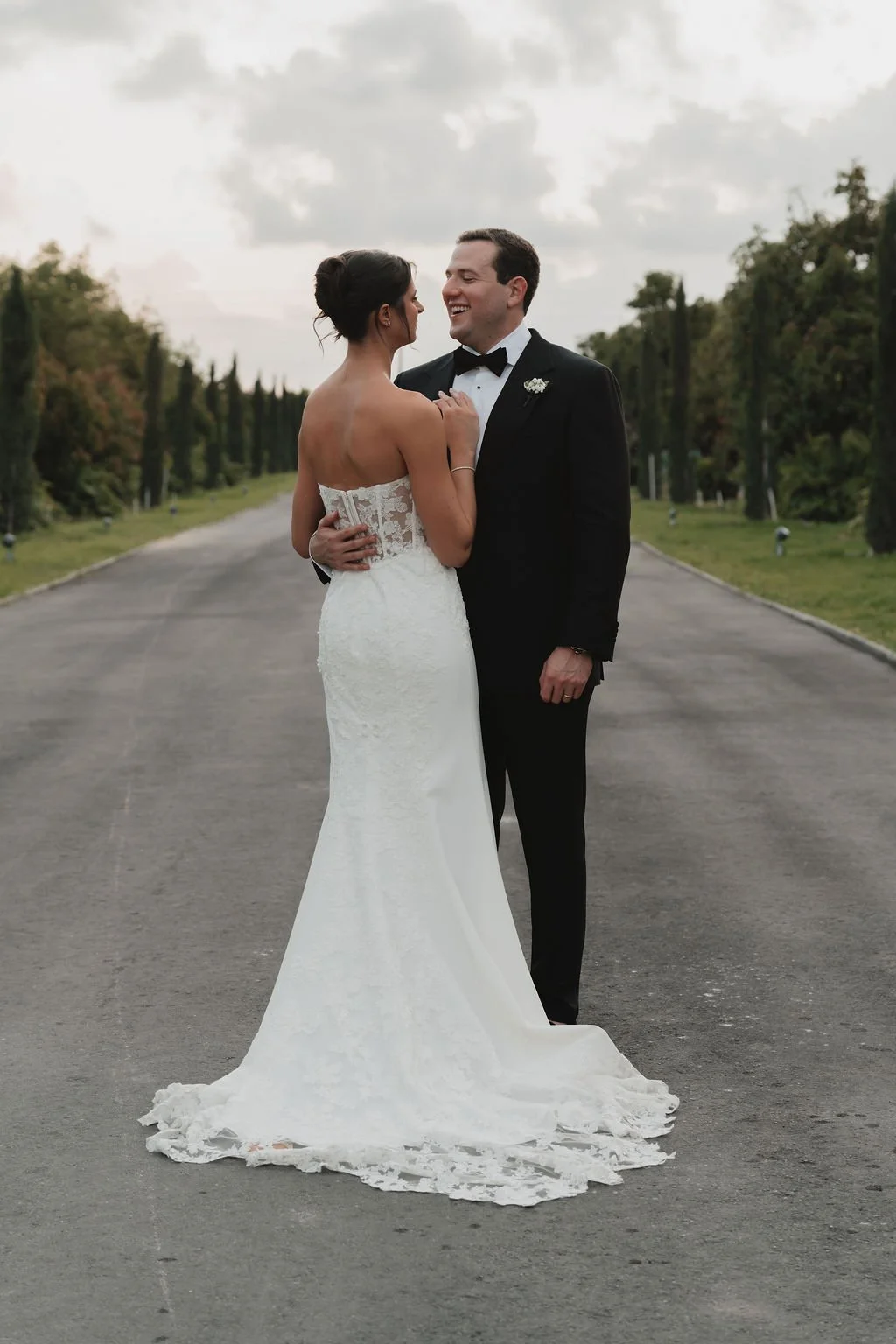 A bride and groom in wedding attire standing on an empty road, embracing and smiling at each other outdoors with trees and cloudy sky in the background.