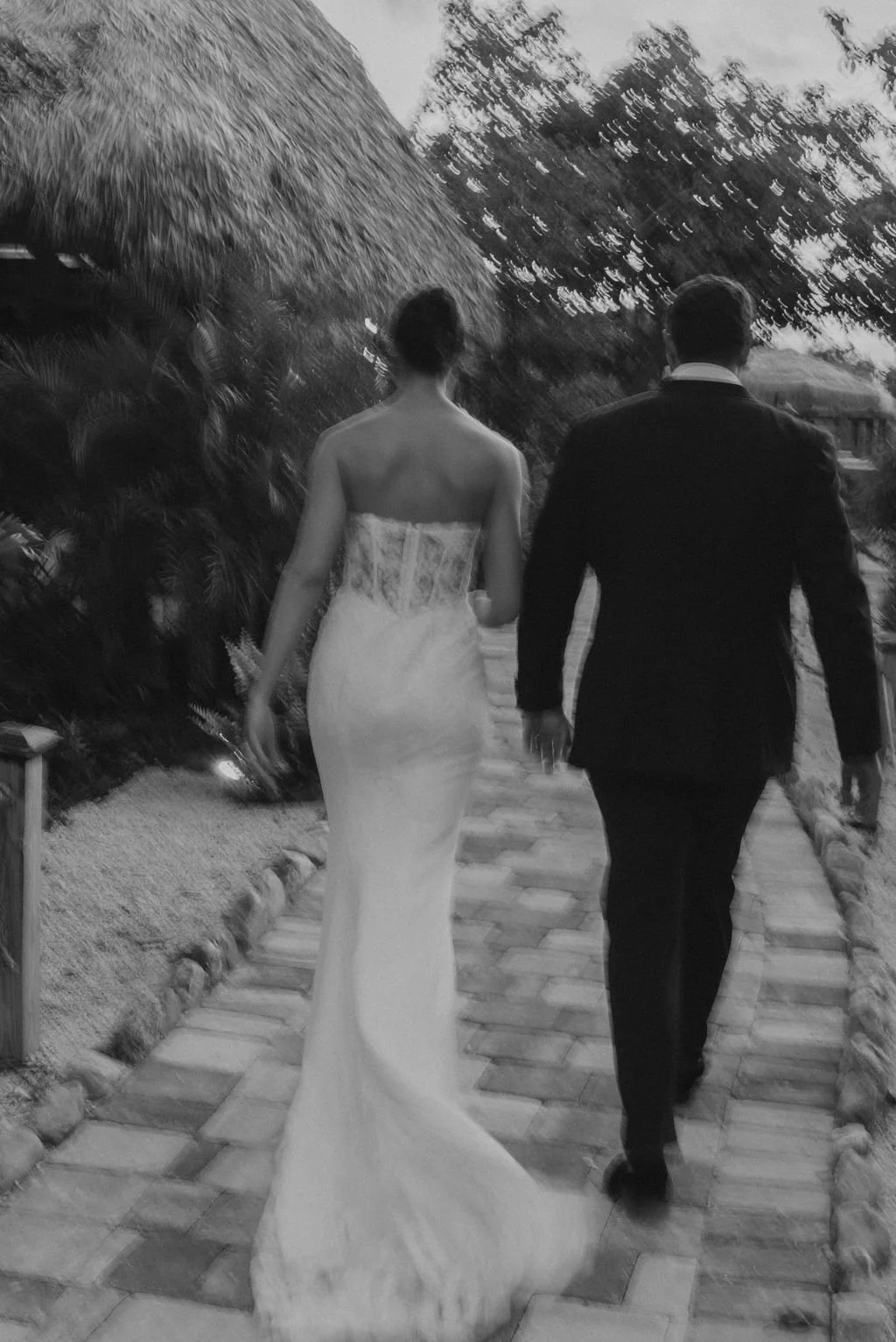 A bride in a strapless wedding gown and a groom in a black suit walk hand-in-hand along a brick path outdoors, surrounded by trees and foliage.