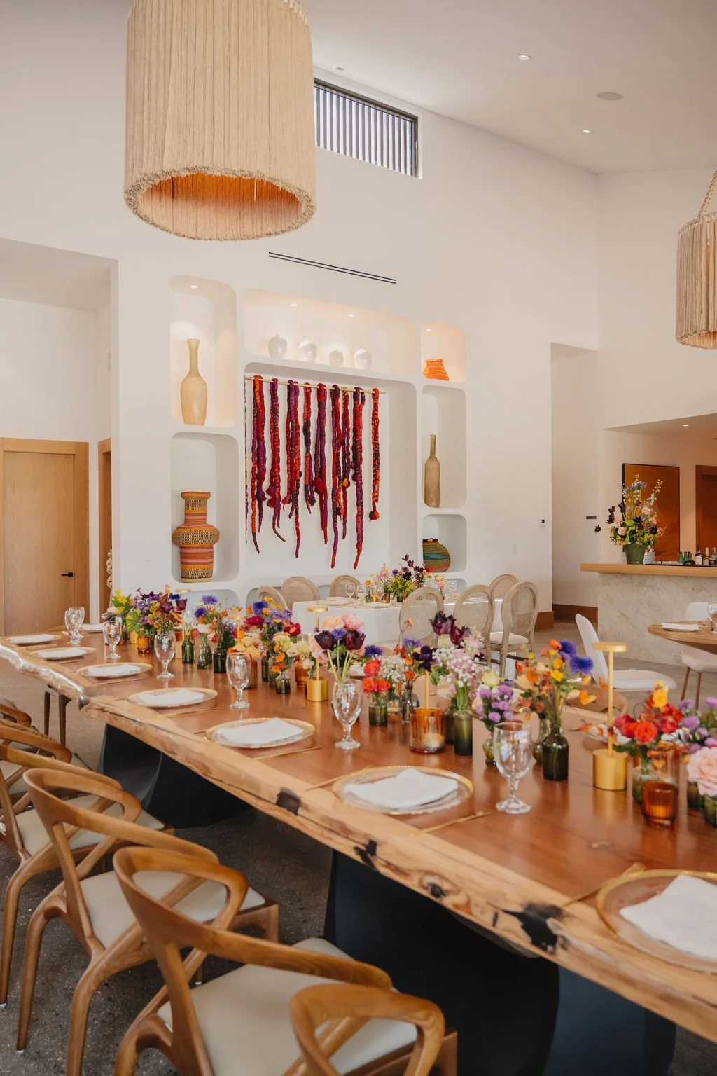 Elegant dining room with a long wooden table set with white plates, glasses, and colorful floral centerpieces. The background features a built-in wall shelf with vases and wall hanging yarn art, and hanging pendant lights.