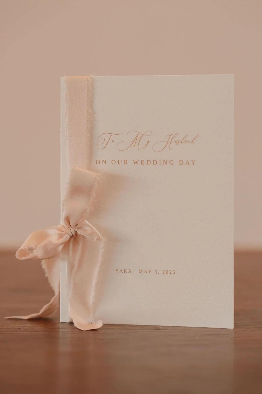 A white wedding card with elegant gold lettering reading 'To My Husband On Our Wedding Day' and 'Sara | May 3, 2025'. The card features a soft pink satin ribbon tied in a bow on the left side.