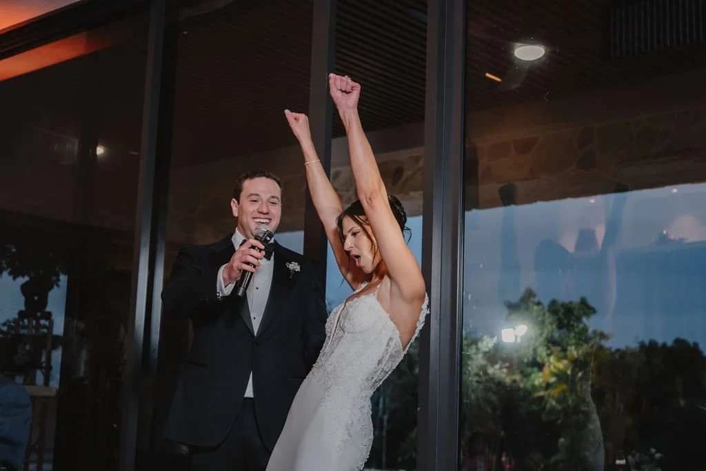 A bride and groom at their wedding celebration, with the bride raising her arm and looking excited, while the groom holds a microphone and smiles.