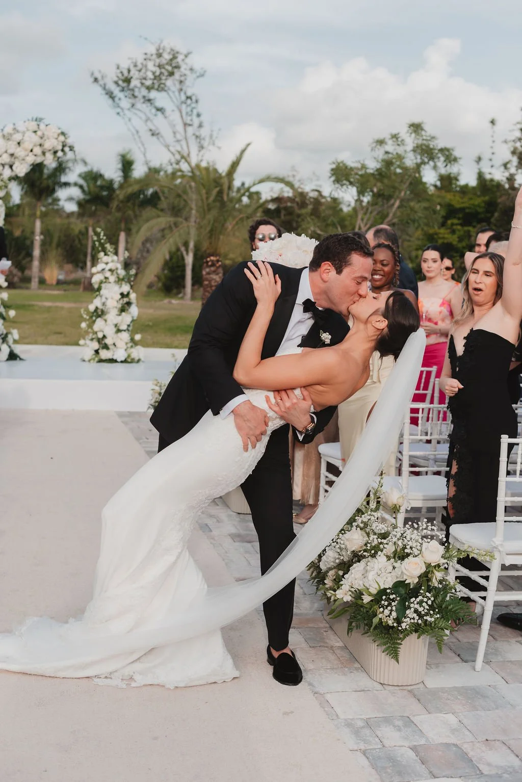A groom and bride share a kiss at their wedding ceremony, with guests happily watching and smiling in an outdoor setting decorated with white flowers and greenery.