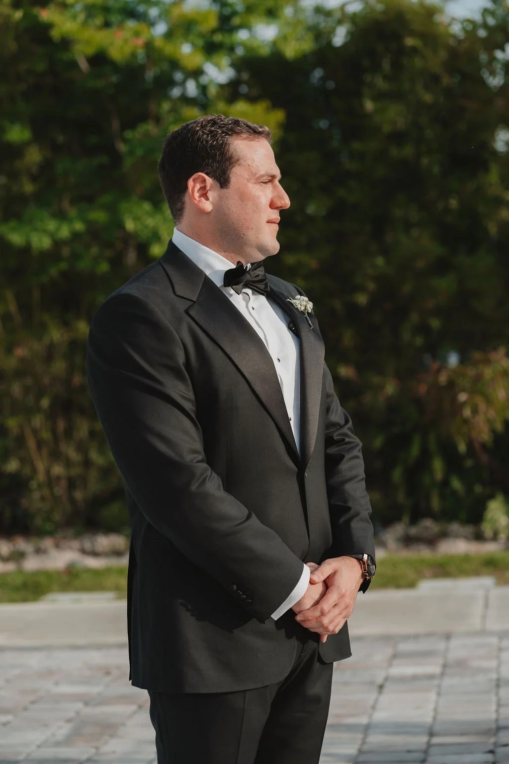 A man in a black tuxedo and bow tie standing outdoors during daytime, with trees in the background.