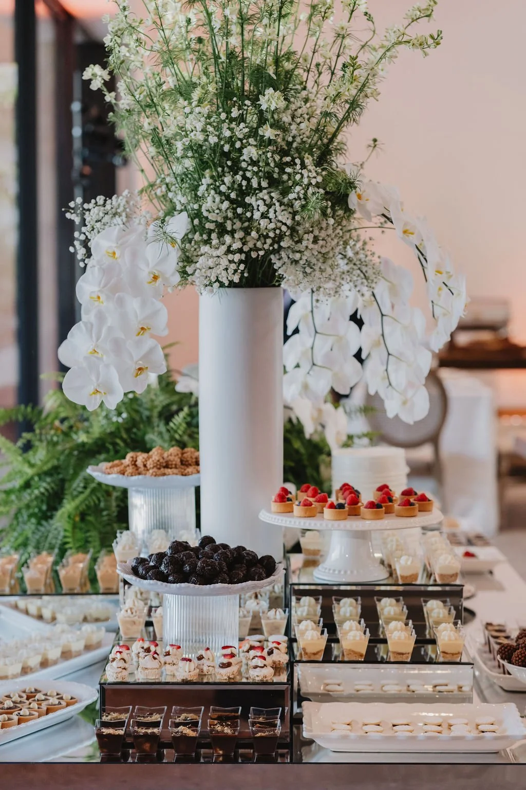 Elegant dessert display with a large floral arrangement featuring white orchids and greenery, surrounded by an assortment of cupcakes, cherries, berries, and other small desserts on white and glass serving platters.
