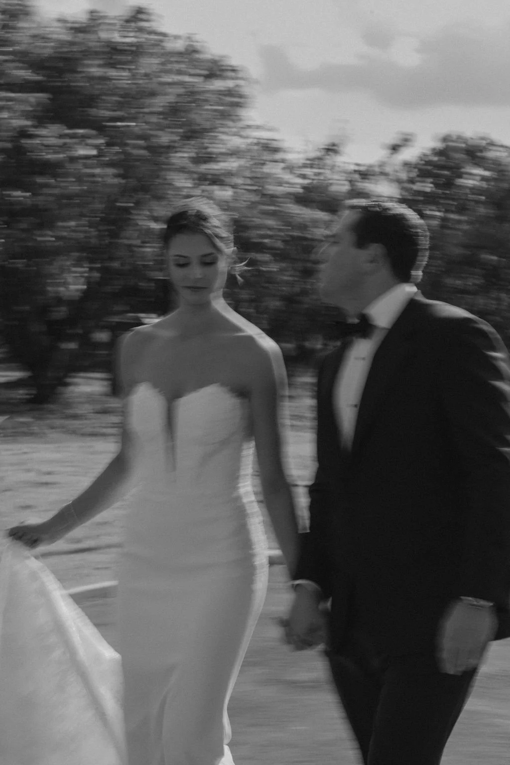 A black-and-white photo of a woman in a strapless wedding dress and a man in a tuxedo walking outdoors, holding hands, with blurred trees in the background.