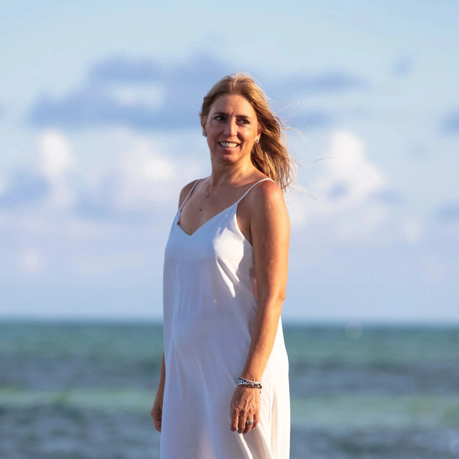 Facilitator, Carina Pucciarelli, woman in a white dress standing on the beach with ocean and cloudy sky in the background.