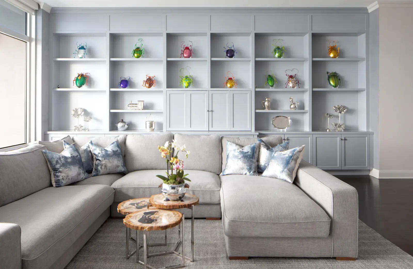 Bright living room with a large gray sectional sofa adorned with silver and blue pillows, a cream area rug, and one potted orchid on a wooden side table. A large white built-in shelves and cabinets filled with colorful beetle sculptures and silver de
