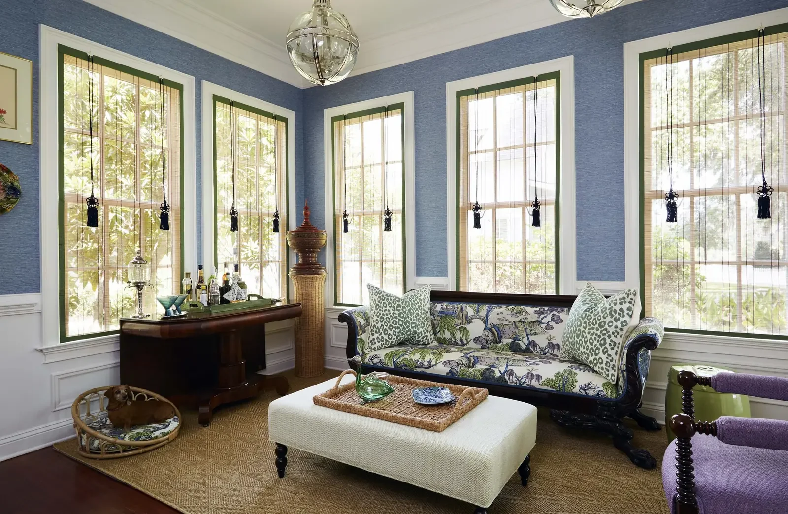 Sunlit living room with blue walls, large windows with black tassel tiebacks, a vintage sofa with floral and animal print cushions, a white ottoman with a woven tray and glass vessel, a wooden sideboard with liquor bottles, decorative lamp, and a pur
