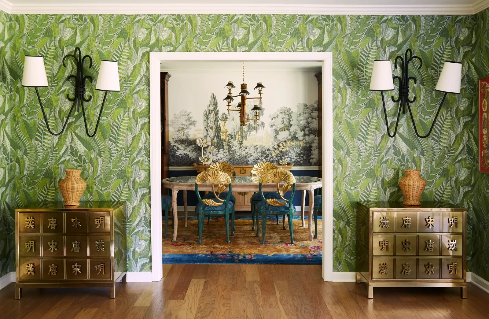 A living room view through a doorway shows two gold-tone cabinets with Asian characters, green leafy patterned wallpaper, black wall sconces with white lampshades, and a dining area with a mural of trees and a chandelier in the background.