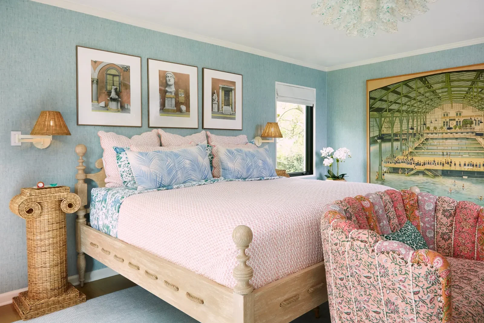 A bedroom with a bed, nightstands, wall lamps, framed art, a window, a large wall mural, and floral patterned furniture and decor.