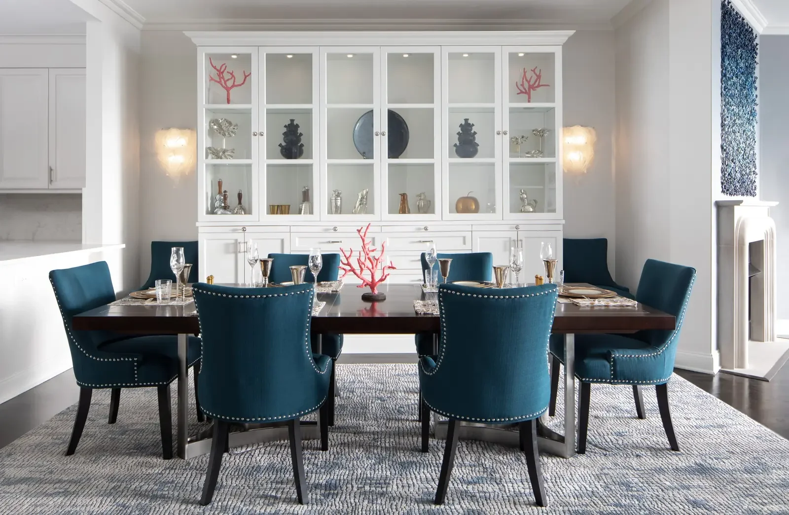 Dining room with a dark wood table and eight teal upholstered chairs with silver nailhead trim, set with glassware and gold-accented tableware, a pink coral centerpiece, and a white built-in cabinet with decorative objects in the background, and two 