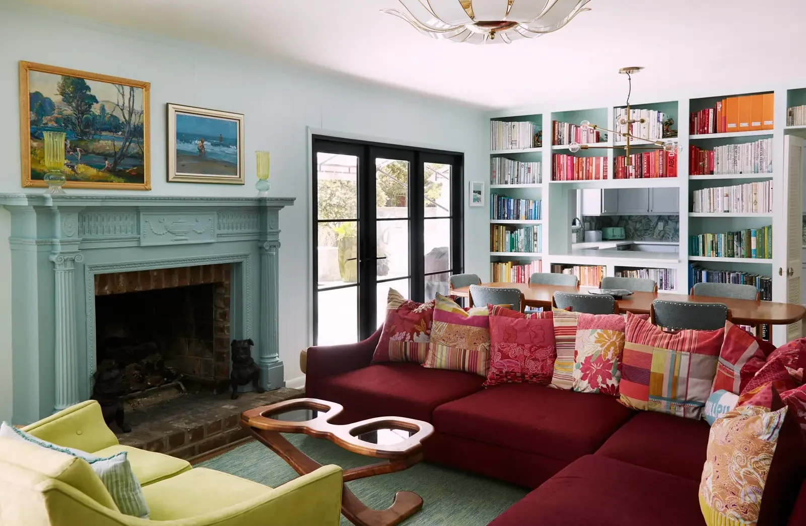 Living room with a fireplace, colorful cushions on a red sectional sofa, a yellow armchair, a wooden coffee table, large glass doors, and built-in bookshelves filled with books.