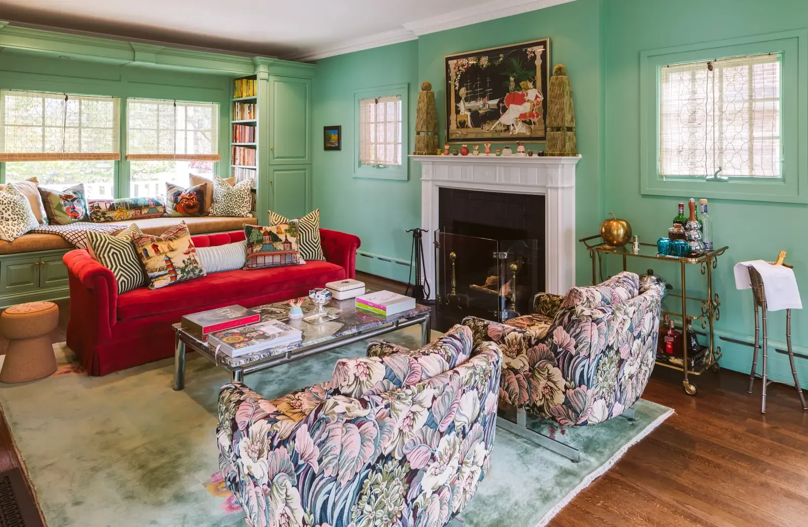 A colorful living room with turquoise walls, a white fireplace, a red velvet sofa with patterned and embroidered pillows, floral armchairs, large windows with floral and animal print pillows, a glass coffee table with books and decorative items, and